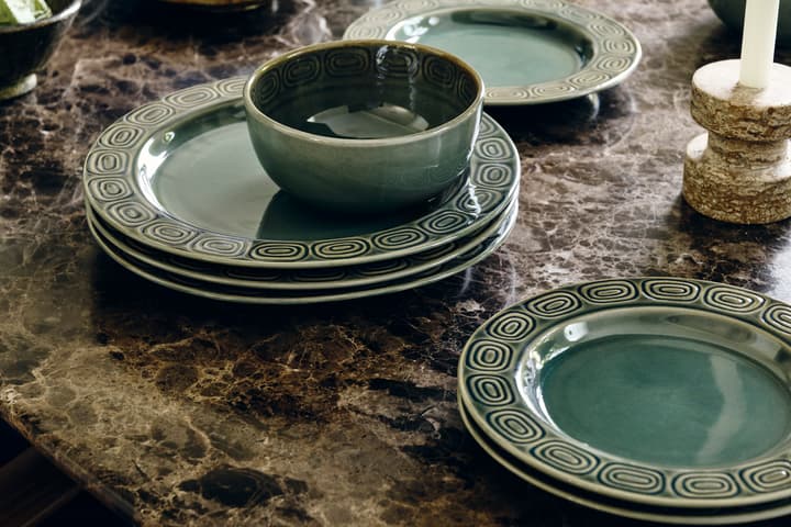 Stack of green, ceramic plates with intricate circular designs, accompanied by a matching bowl, placed on a dark, polished marble tabletop, surrounded by other ceramic dishes and a candle holder.