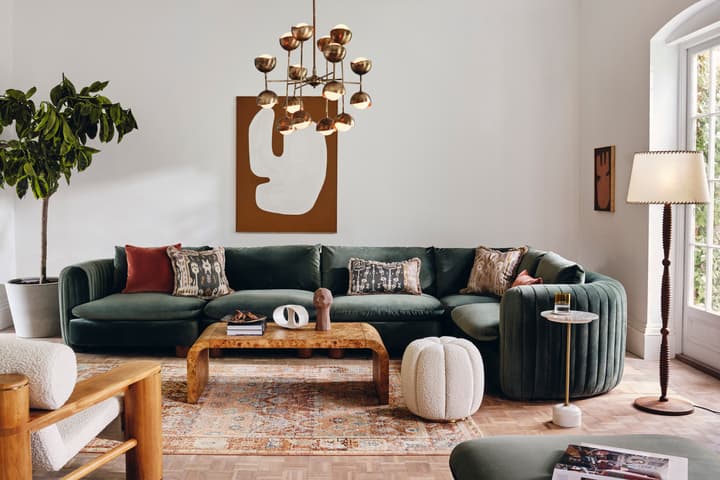 A green sectional sofa adorned with pillows sits against a white wall featuring abstract art, surrounded by a wooden coffee table, a floor lamp, indoor plants, and a textured pouf on a patterned rug.