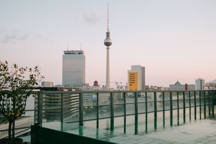 The TV Tower in Berlin stands tall against a pastel sky, alongside the Park Inn hotel and other urban buildings. Foreground includes a rooftop garden and pool area with glass railings. Text on a tall, yellow billboard reads, "You'll never know if you neve