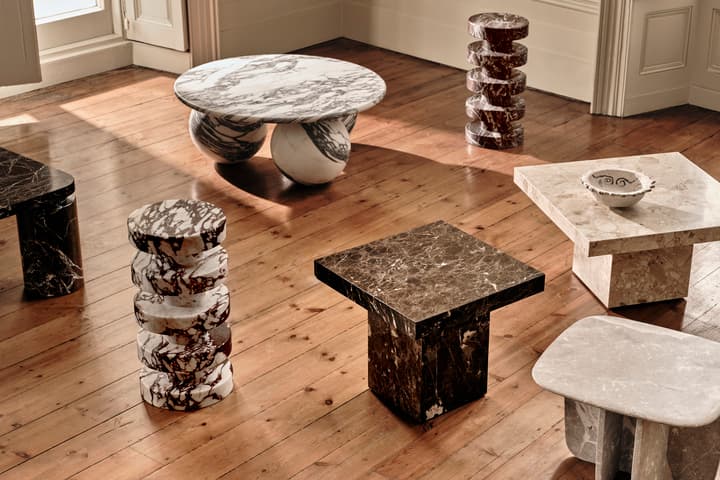 A collection of various marble tables and stools arranged artistically in a sunlit room with wooden floors and white walls. Each piece features different shapes and marble patterns.