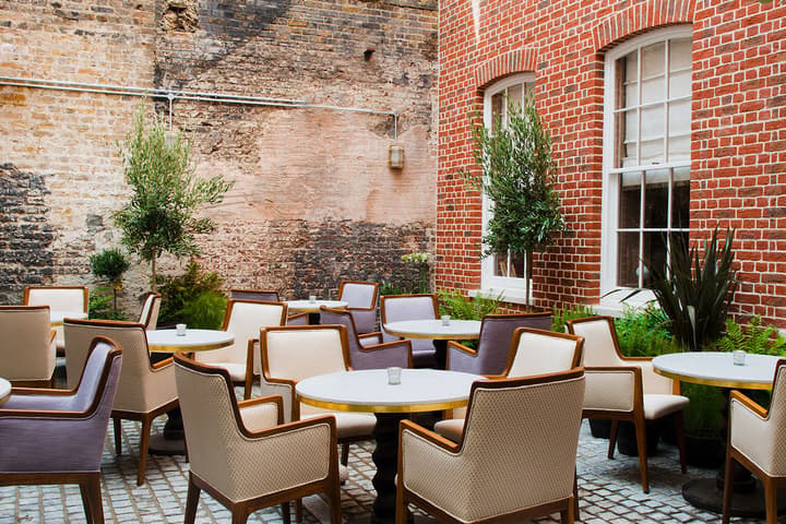 Outdoor café tables and chairs arranged on a cobblestone patio, with a red brick building and an old, weathered mural wall, surrounded by potted plants.