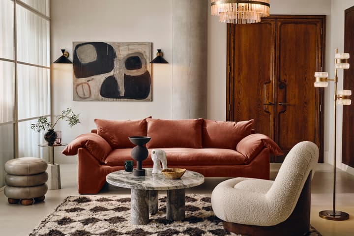 A rust-colored sofa and cream accent chair face a round stone coffee table adorned with decorative items. The room features abstract wall art, warm wooden double doors, and contemporary lighting.