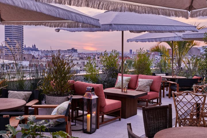 Cozy, outdoor seating with red cushioned chairs and wooden tables, shaded by large fringed umbrellas on a rooftop terrace, overlooks a cityscape at sunset, surrounded by greenery.
