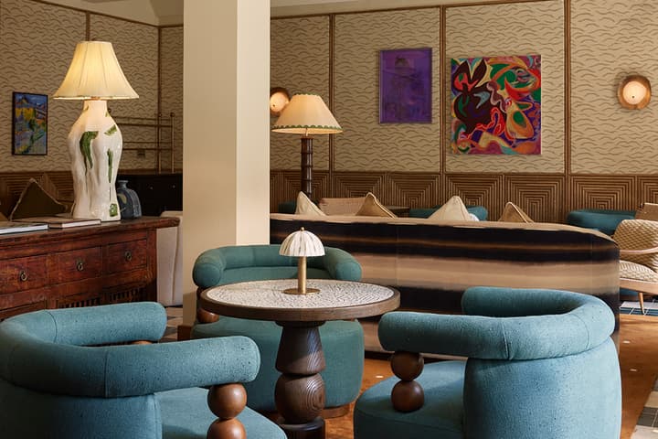 Three turquoise armchairs encircle a round, ornate table with a small lamp, situated in a warmly lit lounge with decorative lighting, abstract wall art, and plush brown carpet.