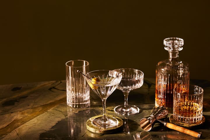 A collection of crystal barware, including glasses and a decanter filled with amber liquid, arranged on a marble tabletop with a brown background.