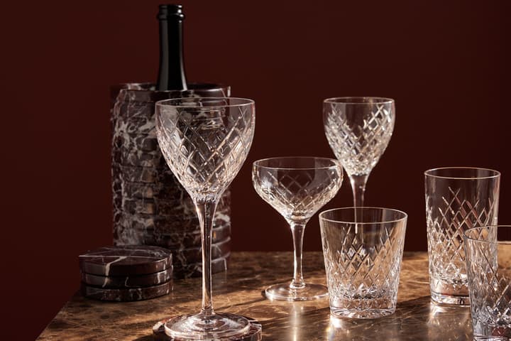 Crystal glassware and a bottle chiller are displayed on a marble table, with a dark maroon background.