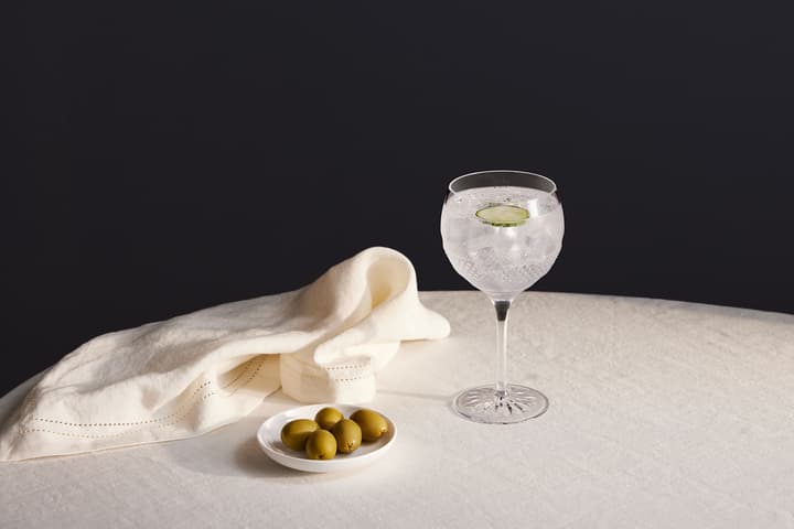 A crystal glass filled with a clear drink and a cucumber slice, next to a folded white napkin and a small dish with green olives, sits on a round tablecloth.