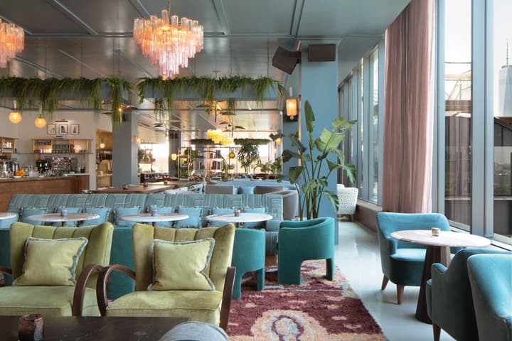 Chairs and tables are arranged in a stylish, modern restaurant with tall windows, chandeliers, and potted plants amid warm, cozy decor. A bar area is visible in the background.