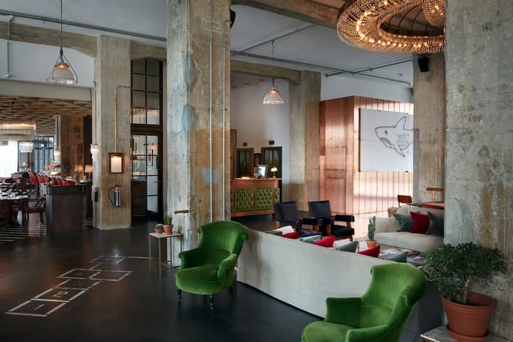 Large industrial-style lounge features concrete columns, green armchairs, sofa with colorful cushions, hopscotch pattern on floor, a crystal chandelier, and wall art of a shark. Background includes a bar and reception desk.