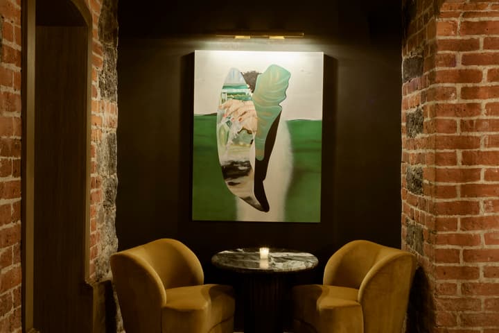 Two yellow velvet chairs and a round black marble table sit against a painted wall with a large abstract artwork, framed by exposed red brick archways and dim lighting.