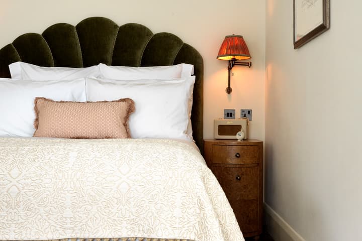 An olive-green upholstered bed with white pillows and decorative bedspread, situated next to a wooden bedside table with a lamp and an alarm clock, in a cozy, light-walled bedroom.