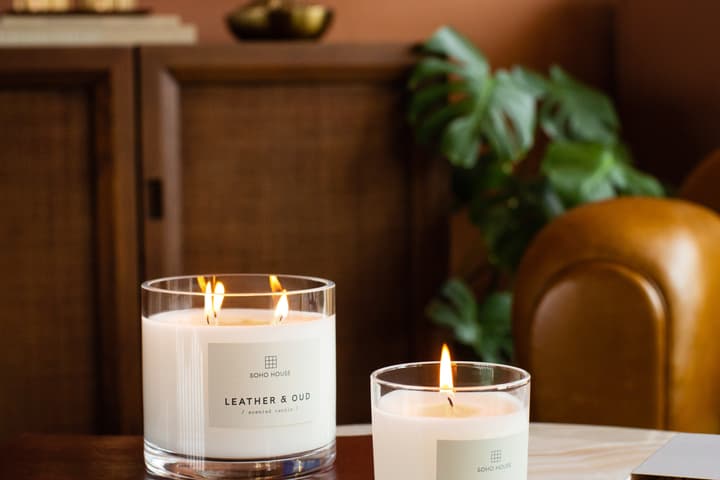 Two lit candles labeled "SOHO HOUSE LEATHER & OUD / scented candle" rest on a wooden and marble surface in a cozy room with wooden furniture and green plants.