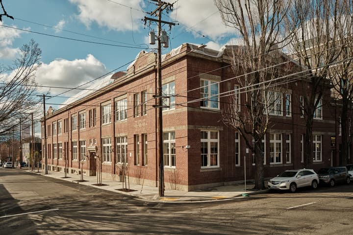 Red-brick building with large windows, situated on a tree-lined street corner; parked cars and utility poles line the sidewalk. The sky is clear with scattered clouds.