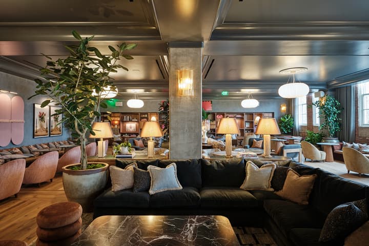 A modern lounge features plush sofas, potted plants, warm lighting from table lamps, with bookshelves and framed art on the walls under a coffered ceiling in an elegant, cozy atmosphere.