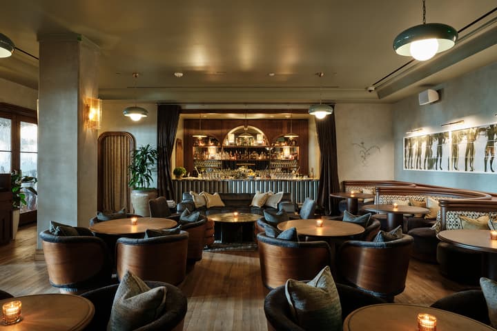A cozy lounge bar features circular tables with cushioned chairs and lit candles; the background shows a well-stocked bar with shelves and decorative lighting in an elegantly designed, dimly-lit room.