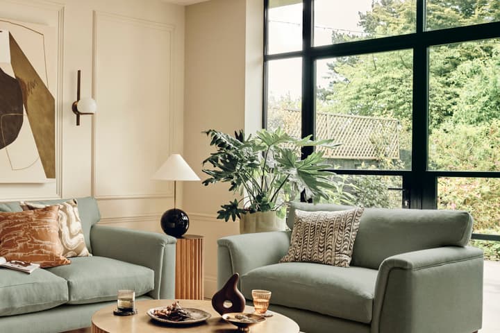 A living room with two green sofas and a round wooden coffee table holding decorative items and glasses. Large windows and indoor plants brighten the space, which includes abstract wall art and warm lighting.