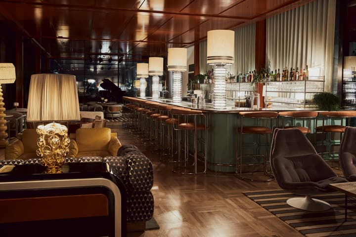 Stylish bar with a long counter lined with high stools, large lamps, and a variety of drinks behind. Chic lounge furniture, warm ambient lighting, and a grand piano in the background.