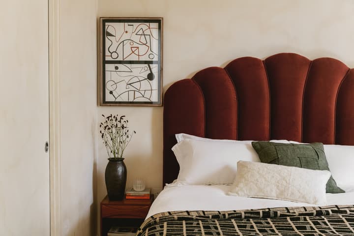 A modern bedroom features a bed with a maroon upholstered headboard, white and green pillows, and a patterned blanket. A wooden side table holds a dark vase with flowers and stacked books.