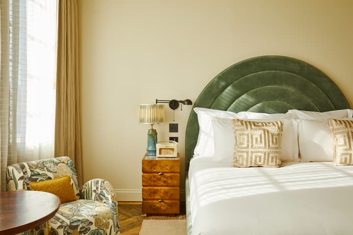 A bed with a green, arching headboard is adjacent to a wooden nightstand holding a table lamp and vintage radio in a sunlit bedroom with a patterned armchair and sheer curtains.