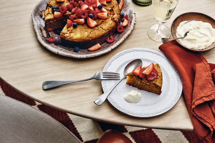 A plate holds a slice of golden cake topped with strawberries beside a dollop of cream, sitting on a wooden table set with utensils, a cloth napkin, and a partial cake.