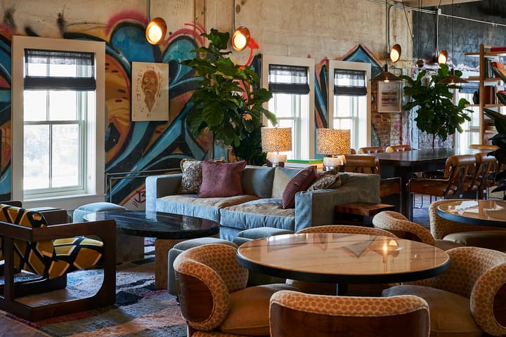 Living room features a gray sofa with colorful pillows, surrounded by art-covered walls, large plants, lamps, and various seating, creating a cozy and eclectic atmosphere.