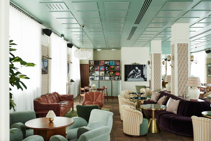 A stylish lounge with colorful modern furniture, including sofas and armchairs, is arranged around small tables. The room features large windows with white curtains, patterned columns, and a display of framed artwork. A shelf filled with vinyl records dec