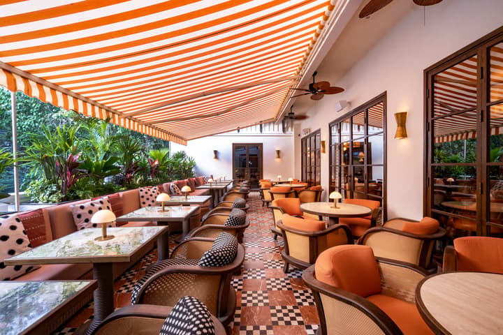 An outdoor seating area features orange-striped awnings, numerous chairs with patterned cushions, marble tables, small lamps, and lush green plants, creating a cozy and vibrant restaurant terrace.