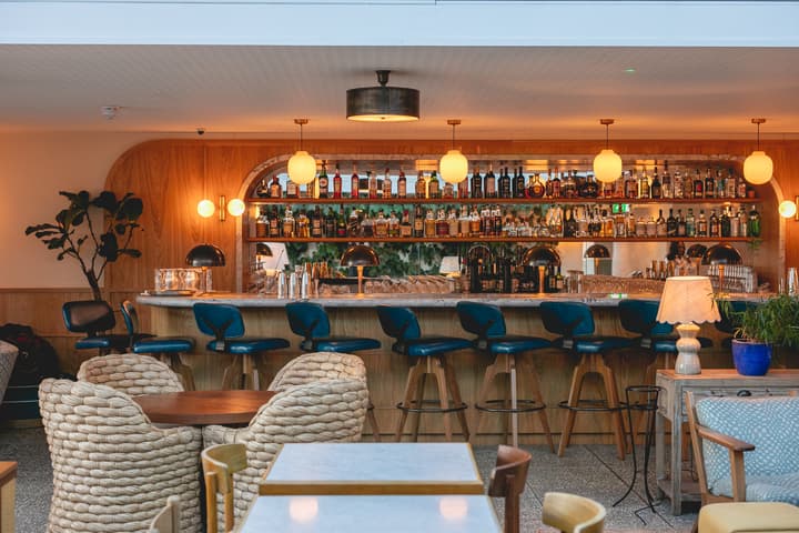 Curved bar counter with blue stools, illuminated by globe lights. Bottles neatly arranged on shelves behind. Wicker chairs and wooden tables in front, plant and lamp on the side.