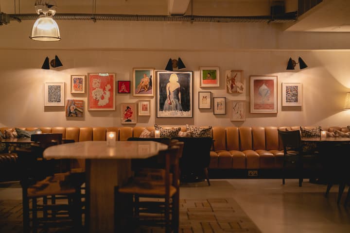 Framed artwork adorns the wall above a tan leather banquette in a dimly lit lounge, featuring wooden tables and chairs, candles, and muted lighting.