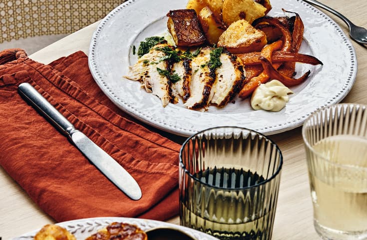 A plate of roasted chicken, potatoes, carrots, and aioli sits on a wooden table set with utensils, a cloth napkin, and glasses of drink, in a cozy dining setting.