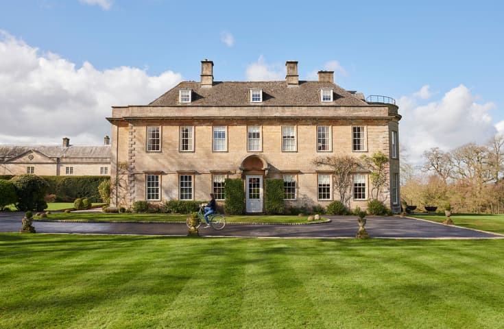 A person rides a bicycle past a large, symmetrical, three-story stone mansion with multiple windows in a landscaped garden with neatly trimmed grass and hedges under a bright blue sky.