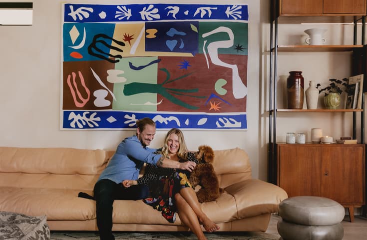 A couple on a beige sofa joyfully plays with a small brown dog in a living room adorned with a colorful abstract painting, a wooden shelf with ceramics, and candles.