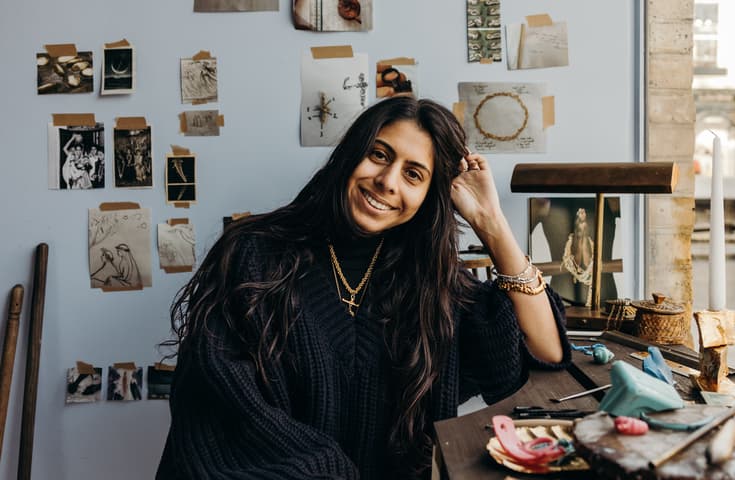 A woman with long hair and a blue sweater sits at a desk, smiling, with artwork and photos on the wall behind her in a well-lit room by a window.