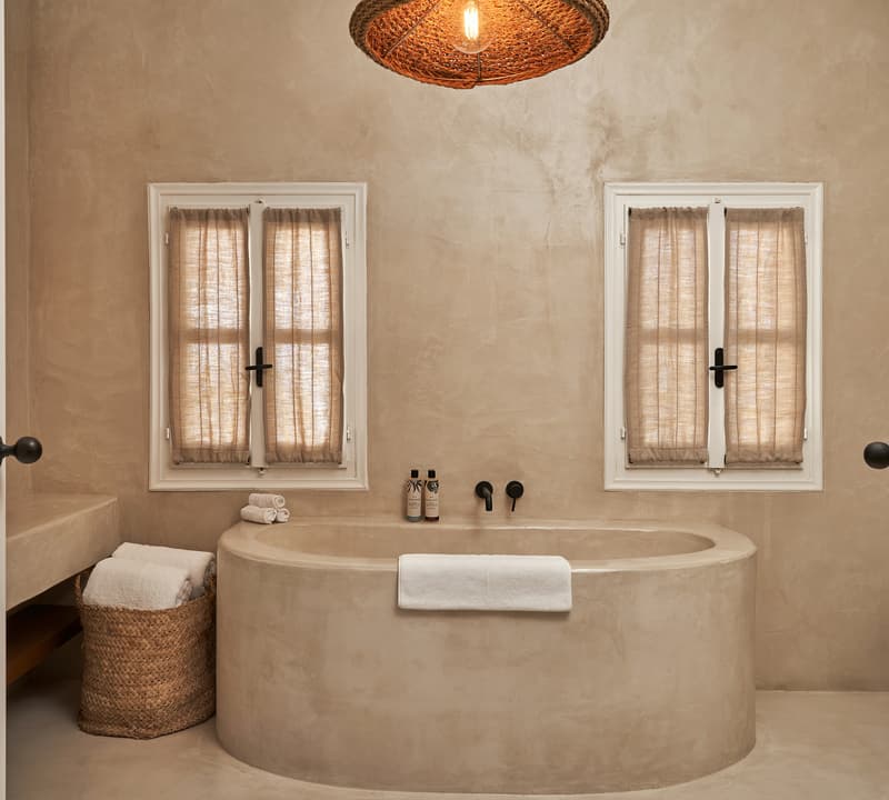 Round bathtub with a white towel draped over it, flanked by two windows with beige curtains, in a beige-toned bathroom. A wicker basket with white towels sits nearby.