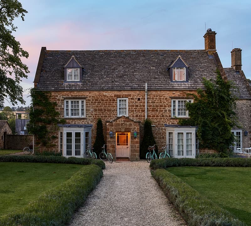 A two-story stone house with ivy on one side, lit warmly from inside, stands among manicured hedges and lawns with several bicycles near the entrance at dusk.