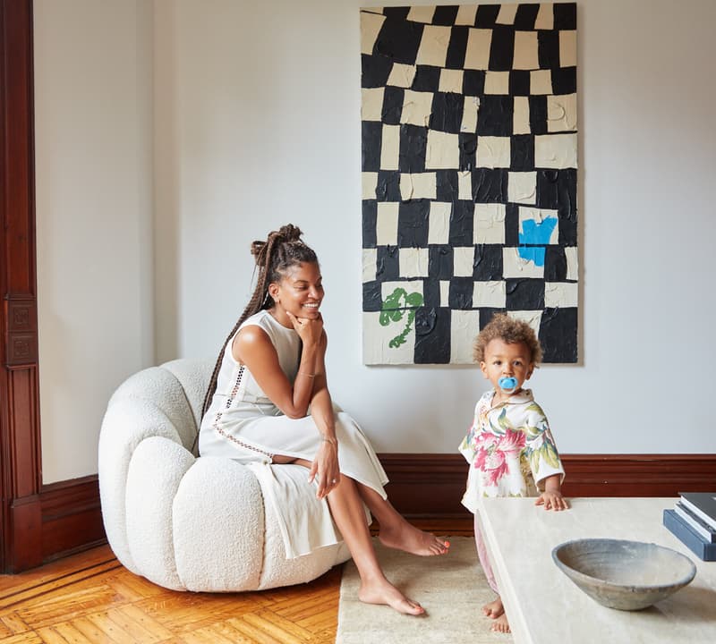 A woman sits on a stylish white chair, smiling, while a toddler with a pacifier stands nearby by a coffee table in a cozy, well-lit living room with an abstract checkerboard painting on the wall.