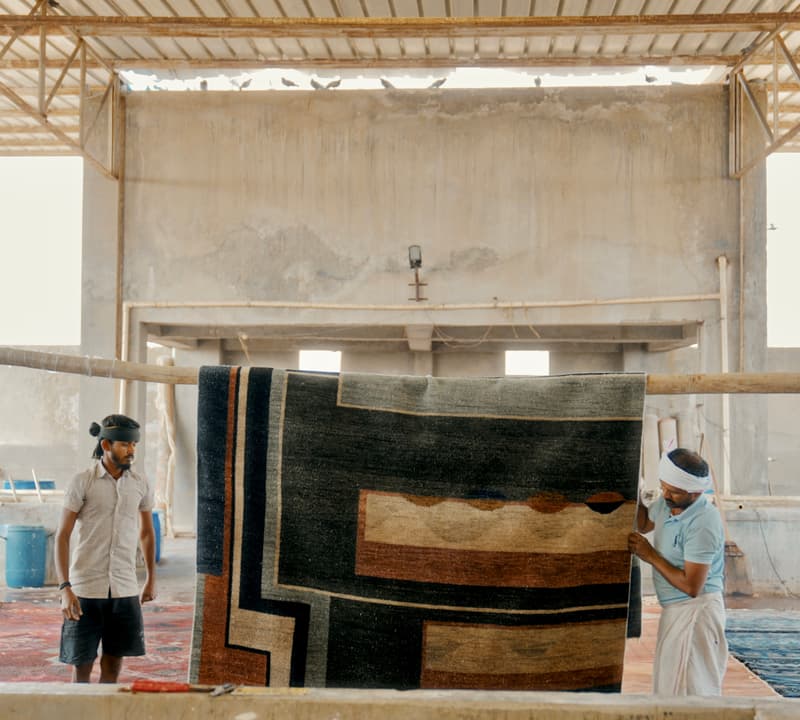 Two men hang a large geometric-patterned rug on a wooden beam, inside a spacious, industrial-looking room with a high, metal-framed roof.