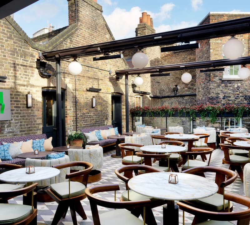 Circular marble tables and cushioned wooden chairs arranged in a cozy outdoor patio, surrounded by brick walls adorned with greenery and modern lights, create a relaxed and inviting atmosphere.