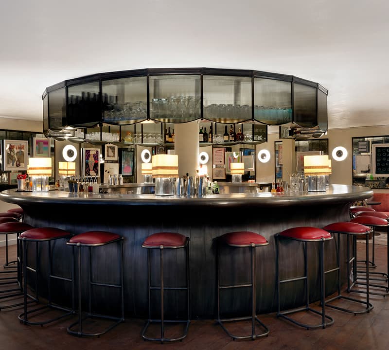 A circular bar with red stools is surrounded by mirrors and shelves holding bottles and glassware. It is lit by modern lamps, and the walls display various framed art pieces.