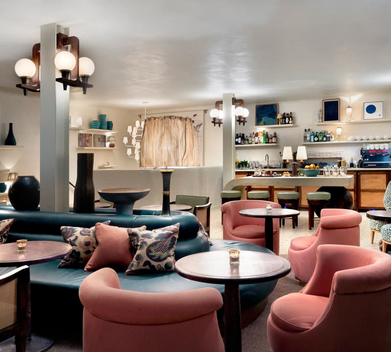 Lounge area featuring plush, pink and blue chairs and a sofa around small tables. Background includes a fully stocked bar, table lamps, wall art, shelves, and warm, ambient lighting.