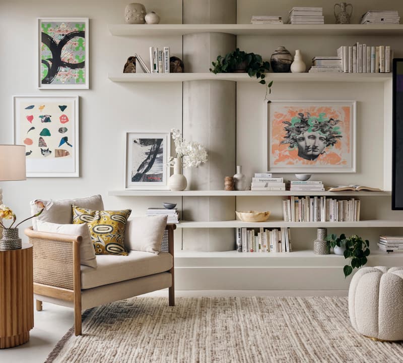 A cozy, modern living room features a cushioned armchair beside a wooden side table with a lamp and flowers. Art prints and books adorn the wall shelves, complementing the neutral-toned decor.