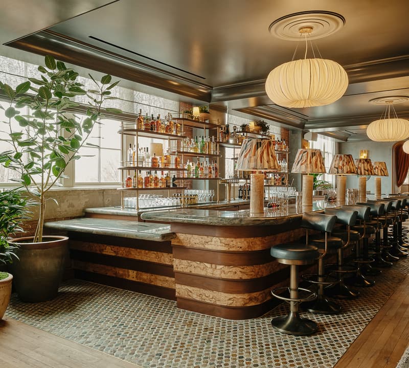 A well-lit, stylish bar featuring a marble countertop and cushioned stools, with shelves of liquor bottles. Potted plants adorn the room, which has modern light fixtures and a wooden floor.