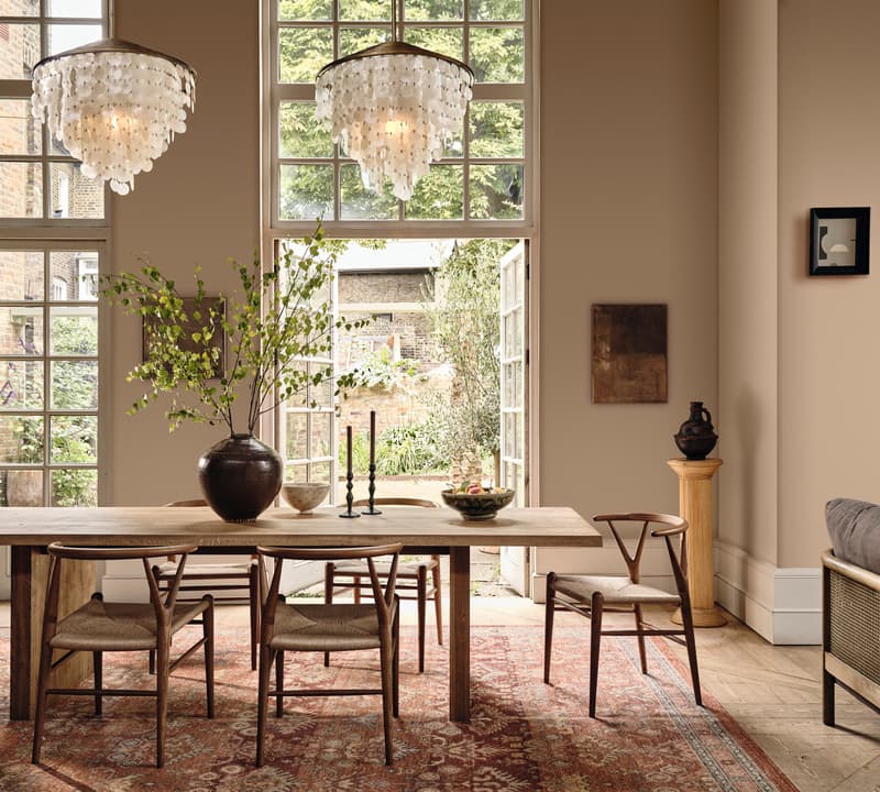 Dining table with wooden chairs and a vase of branches, set under two chandeliers. Context: spacious room with tall windows, beige walls, a red-patterned rug, and visible garden outside.