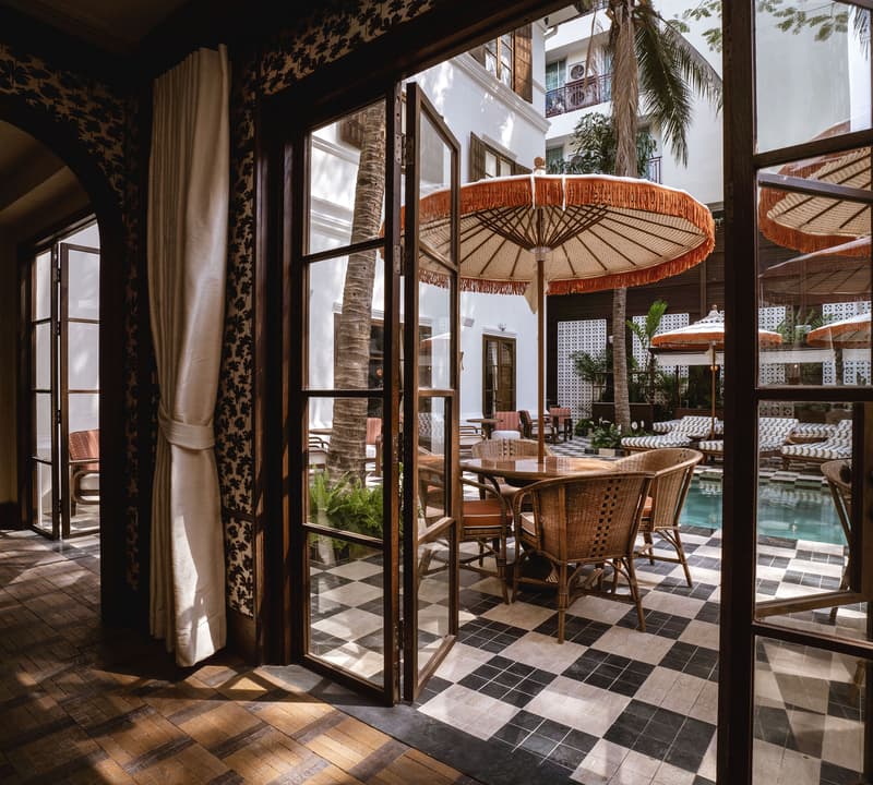 Open double doors lead to a stylish patio where wicker chairs surround a table under large, fringed umbrellas beside a swimming pool in a tropical courtyard with palm trees.