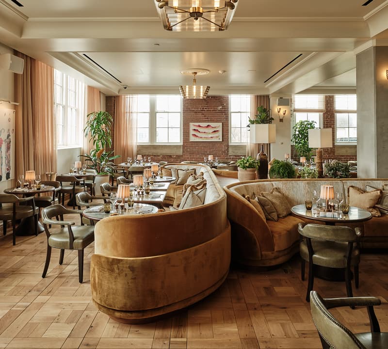 Elegant, well-lit restaurant with plush tan booths, round tables set with lamps and tableware, surrounded by large windows, brick walls, and abundant greenery. Soft beige drapes and tasteful art adorn the walls.