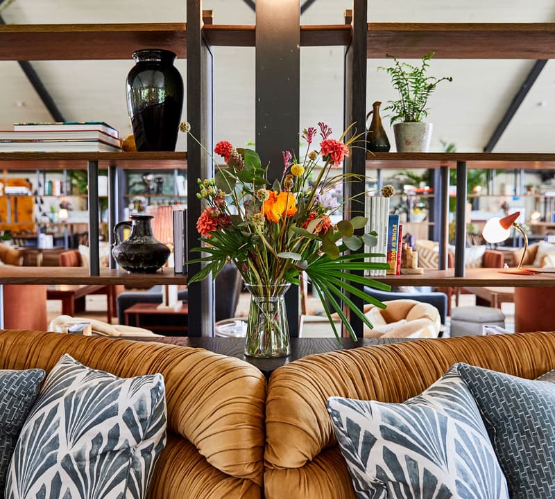 A vase with colorful flowers adorns a shelf. Surrounding objects include various books, vases, lamps, and plants. The background reveals a cozy, stylish, and well-decorated living space.