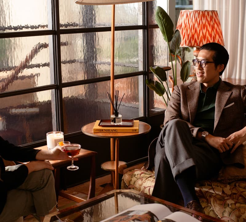 Two people converse in a cozy, well-lit living room with a patterned couch, large windows, a lamp, plants, and a book titled "DUNHUANG" on a side table.
