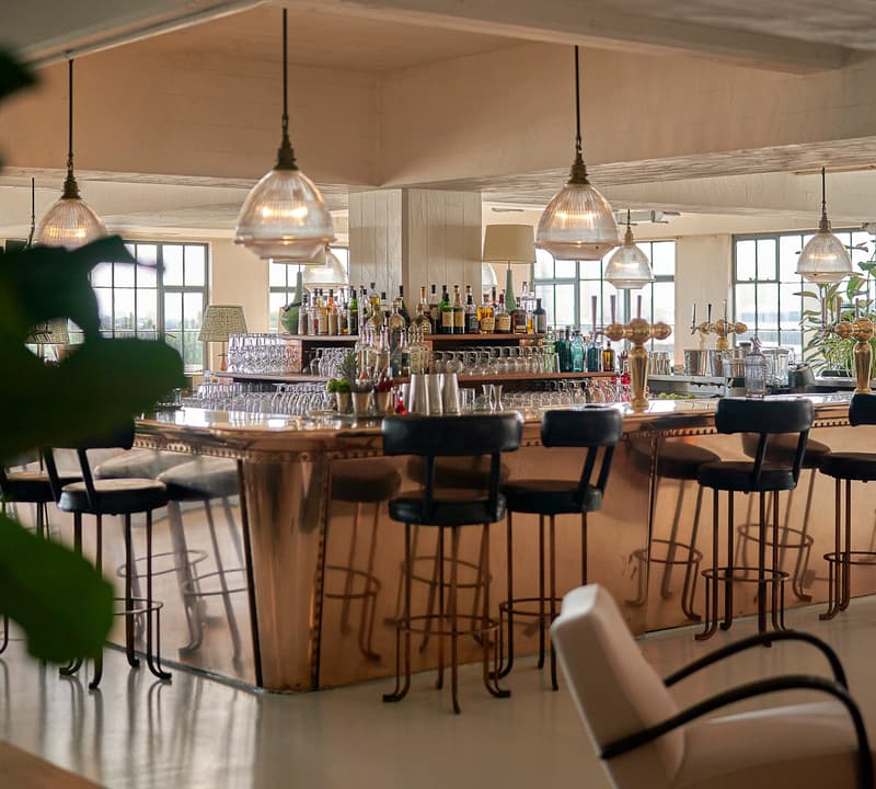 A stylish bar with a polished copper counter, surrounded by black cushioned barstools and under warm pendant lights, sits in a spacious, well-lit room with large windows and modern decor.