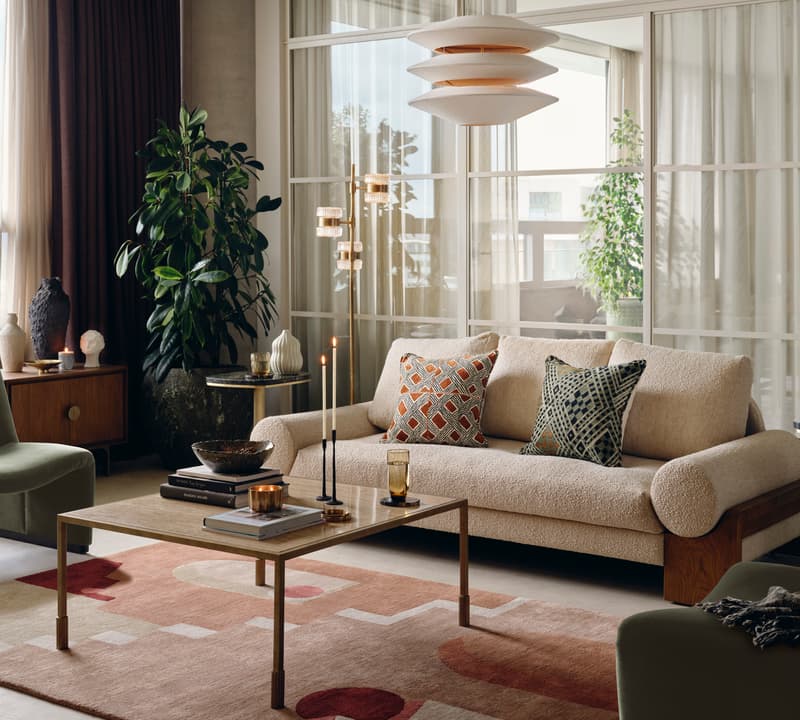 A contemporary living room featuring a beige sofa with patterned cushions, a coffee table with candles and books, adjacent to armchairs, houseplants, and modern decor, illuminated by natural light and an overhead lamp.