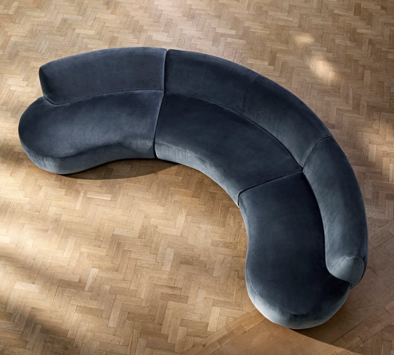A curved, gray sofa sits empty on a light herringbone wood floor, viewed from above, with no other furniture or decor visible in the room.
